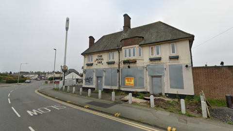 The boarded up pub stands on a corner of a road. It is beige in colour with metal covers on lower ground windows. There are small bollards outside it on the bend in the road.