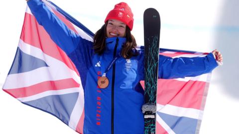 A woman in blue Team GB ski suit holds a union jack up behind her back between her outstretched hands. She is wearing a bronze medal and red beanie hat and has a black ski leaning on her left shoulder.