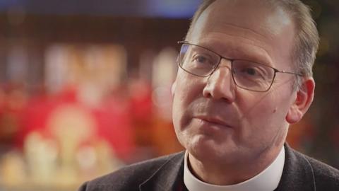 Head and shoulders image of Peter Howell-Jones wearing a dog collar and black jacket. He has short fair hair, greying at the sides, and black wire-rimmed glasses. In the background are blurred candles