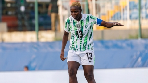 Ngozi Okobi-Okeoghene sports short, dyed-blonde hair while wearing a white and green Nigeria football shirt and white shorts with the number 13 visible on both. Her left arm is extended to her side for balance as she looks down intently during a football match. Blurred in the background are fences positioned in front of a stand with seats in it