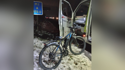 An e-bike on the snow-covered hard shoulder of the M1 in Leicestershire leaning on the open door of a police van