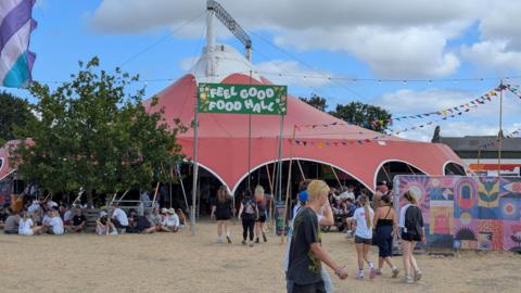 People walking by the Feel Good Food Hall tent at Truck Festival. There is a group sitting beside it. There is a colourful mural on the right. It is cloudy.