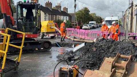 A road with a large square hole in it. Workers in orange hi-visibility clothes can be seen crowded around it. There are construction vehicles and equipment at the scene. 