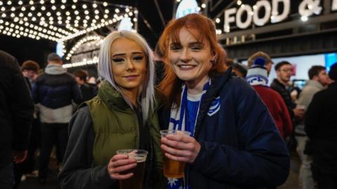 Two women, one with ginger hair and one with white blonde hair, holding pings of beer and smiling at the camera. There's a food stall and some fairy lights behind them, as well as crowds of people.