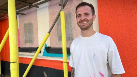 A man in his late 20s wearing a white T-shirt with pink and blue paint streaks on it smiles at the camera. He has short brown hair and brown stubble, and has tattoos poking out of his sleeves. He is standing in front of an external wall with different colours of paint on it, but is mostly bright orange, with two spaces painted to look like windows especially for theatre posters.