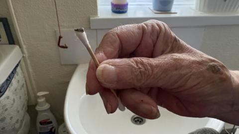 A hand in a bathroom holding a cotton bud covered in dark sludge. Behind is a sink and some cleaning products.