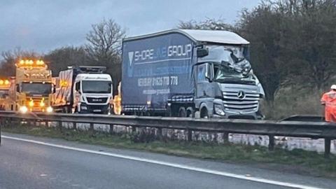 A large blue lorry with crumpled cab and roof, a white lorry is pulled up behind it along with several bright yellow recovery lorries with lights flashing.