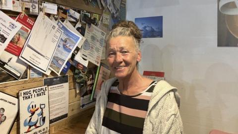 Amanda Wilkinson has greying brown hair in a bun and wears a stripy top of different shades of brown with a light hoodie.