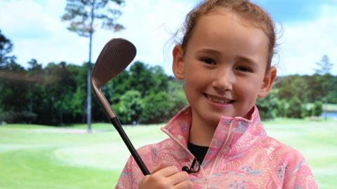 Hope in a pink zip-up sweatshirt. She is looking at the camera and is smiling while holding a golf club. She is stood in front of a screen that has a golf range on it.