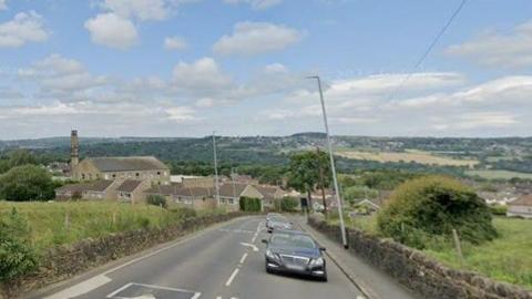 A road with a dry stone wall on either side, surrounded by fields. In the backdrop are houses, buildings and hills. There are three cars shown being driven on the road.