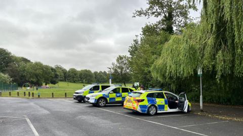 Police cars in a car park, with a green park lined with trees behind them. A play area is to the left.