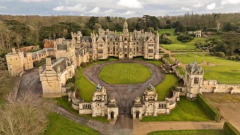 An aerial view of Harlaxton Manor in Lincolnshire. The house is surrounded by a large wall featuring several towers. A circular lawn is situated in the centre of a courtyard in front of the main building. Landscaped gardens are on the right of the image.