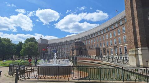 Entrance to city hall which has lots of windows. There is a fountain outside, near to a grass area.