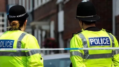 Two police officers stand with their backs to the camera in uniform with fluorescent jackets on and helmets. Police tape is also visible behind them. They are standing in a street looking towards a building that is blurred out.