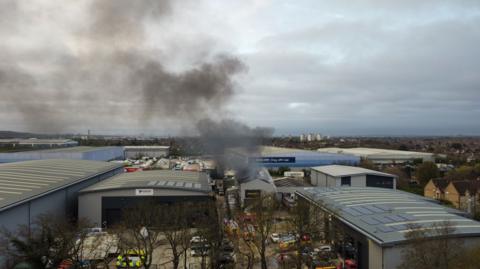 An aerial shot of the industrial estate, showing smoke coming from the roof of one building. There are many large warehouses around and emergency vehicles next to it.