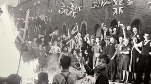 People laughing and smiling while celebrating VE Day in a London Street that has been bombed. There is a bonfire in the street and there is Union Jack bunting above their heads and a child with his face turned to the camera is waving a flag on a pole.