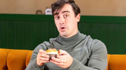 An actor resembling Joey from Friends pulls a face and holds a cup cake while sat an an orange sofa reminiscent of Central Perk