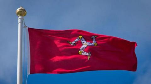 The Manx flag flying at full mast in the wind, it is a red flag, with a white triskellion in the middle and golden knees and feet.