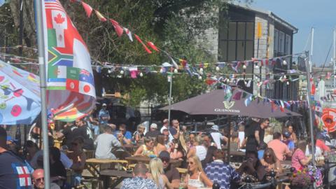 A picture of a number of people sat in a pub garden. There is a number of flags pictured above the tables.