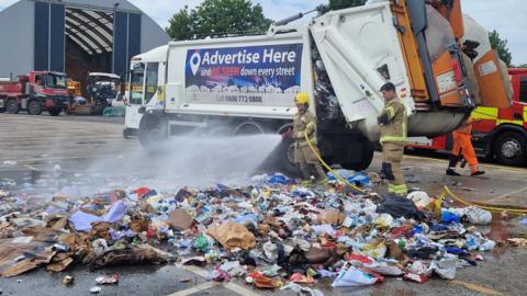 Fire crews hosing down rubbish in a pile near to a bin lorry in a depot