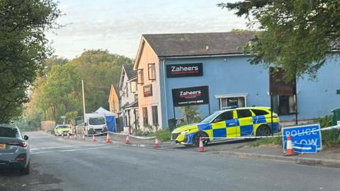Two police cars and police tape around a row of three buildings next to a road. 