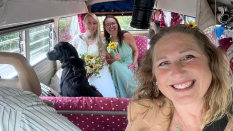 A selfie taken by Cat Lawson, Paige Lawson's mother, shows her in a campervan with bride Paige Lawson and her bridesmaid on a seat in the background, while a dog sits looking out of the window 