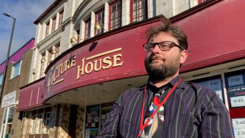 A man in a multi-coloured tie and striped shirt standing outside an historic cinema.