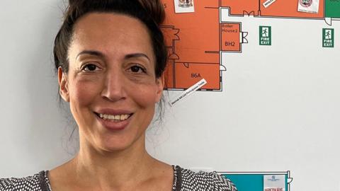 Nicola Welham is the centre manager of The Charles Burrell Centre a former high schools turned into community and business hub. She is wearing a black and white dress and has her brown hair in a bun. She is standing next to a map of the centre