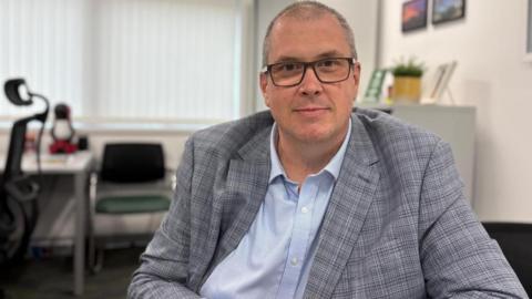 A bald man looking at the camera in an office. He has a light blue shirt and a grey checkered jacket