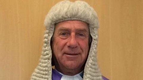 Judge Martyn Levett smiles at the camera as he stands in front of a wooden wall. He wears a judge's white wig, purple robe and white shirt. 