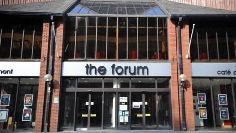 A general view of entrance to The Forum in Barrow, Cumbria. The signs above the glass doors and windows are black and white. There are show posters on the windows.