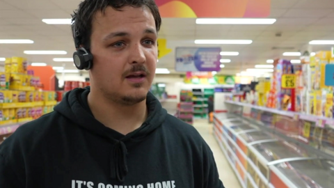 A person wearing a black “IT’S COMING HOME” hoodie and a headset stands in the frozen food aisle of a brightly lit supermarket, with freezers.