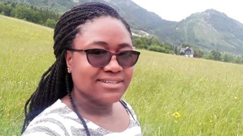 A young black woman wearing sunglasses with her hair in braids, posing for a selfie in a meadow with hills in the background