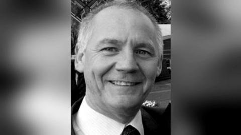 A black and white image of Paul Taylor. He has balding grey hair and is smiling at the camera, wearing a suit.