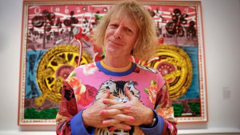 Grayson Perry folds his hands while posing in front of his painting