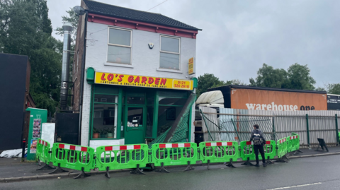 Green barriers surround a takeaway called Lo's Garden which is a detached building on the street. It has a green frontage and yellow sign with a floor above the takeaway. Part of a metal covering across the lower floor of the takeaway is hanging off over the pavement which the barriers are protecting. A man is standing by the barrier looking at the shop.