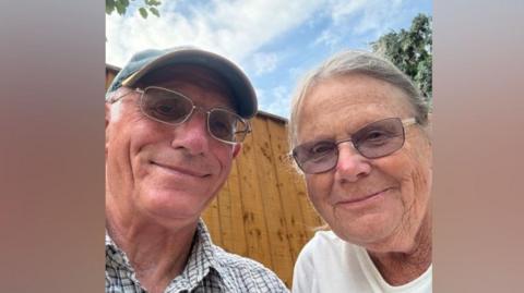 A selfie-style image of an elderly man and an elderly woman. They are wearing glasses and smiling while looking into the camera. The man is wearing a baseball cap. They are both wearing glasses.
