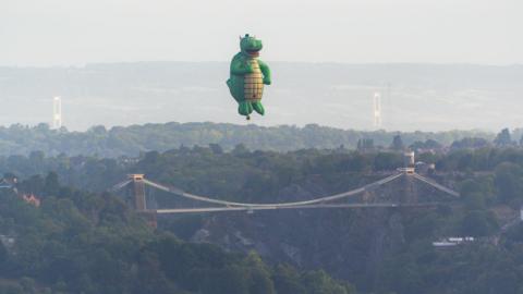 A giant green hot air balloon in the shape of a dragon flies high above Clifton Suspension Bridge. It has a yellow belly and a smiling mouth as well as two small horns. The Prince of Wales bridge is just about visible in the far distance.