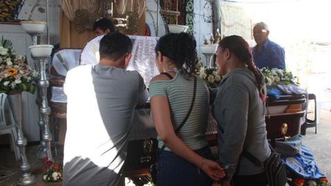 Two women and a man gaze at the open coffin of one of the victims of the shooting in Playas, Ecuador. Flowers have been placed on the closed part of the lid and the white lining of the coffin can be made out where it is open. In the background, there is a second coffin. 