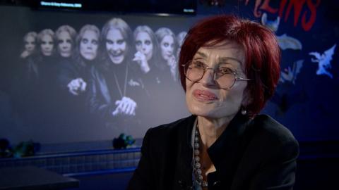 A woman is smiling off camera. Sharon Osbourne has red hair, thin rimmed glasses and red lips. Also wearing a black jacket and pearl necklace.