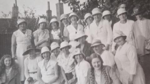 A black and white image of a group of women wearing white bowler hats and white dresses. They are smiling at the camera. 
