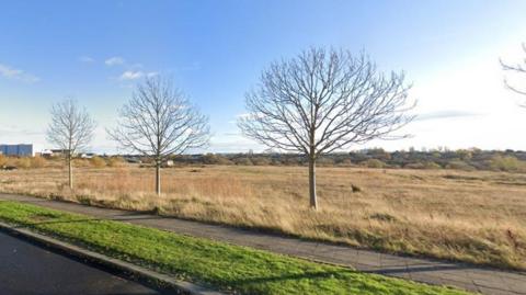 The large land off Cygnet Drive in Stockton is surrounded by several buildings in the distance. A number of young trees a re growing between the grassland and the pavement.