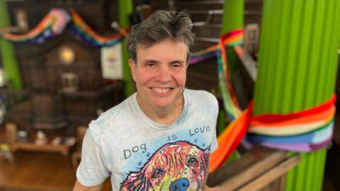 Helen Simpson, a woman with short grey hair, is looking into the camera, smiling. Behind her, there are green pillars, and the pride scarf, which wraps its way around the room.