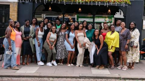 A group of 21 young black women on the street outside a restaurant smiling and posing for a picture