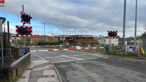 Level crossing at Ash is closed, with red lights. Pavement in view and a building with scaffolding in the background.