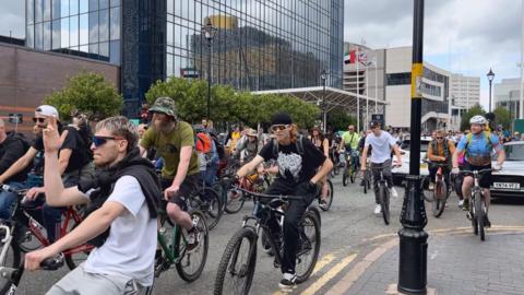 A group of men are cycling on their bikes, some are wearing helmets and glasses. They are dancing along to music as they cycle on a road. 