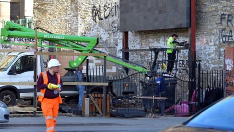 Construction workers and equipment at the site. One person walks away from the site in orange high-vis with a bag of crisps in his hands. Another person carries out some work while standing on a cherry picker