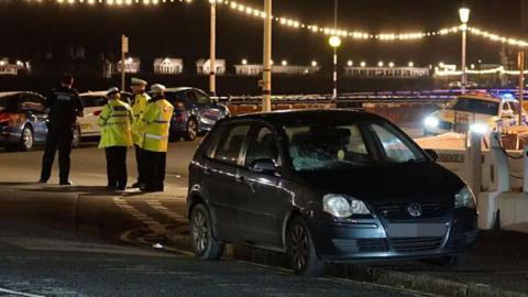 A dark car with police officers in hi vis jackets behind it and police tape on the seafront in Eastbourne at night 