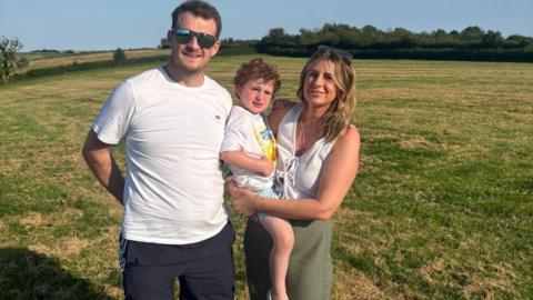 Richard stands with his family in a field on a sunny day. He has short dark hair and reflective sunglasses and a white t-shirt. A woman with blonde hair, her sunglasses on her head smiles beside him. She is holding a boy on her hip, with Richard leaning into his back. The boy has curly red hair.