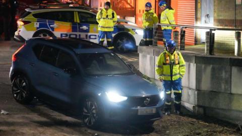 A car with its headlights on on a ramp near the river. Four people in high-visibility jackets are stood nearby. A police car is in the background.